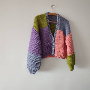 Op de afbeelding: Een chunky gebreid vest in een patchwork-ontwerp, met panelen in lavendel, lichtblauw, roze, groen en olijfgroen. De cropped trui heeft een V-hals, knoopsluiting en lange mouwen. Het hangt aan een houten hanger tegen een witte muur.