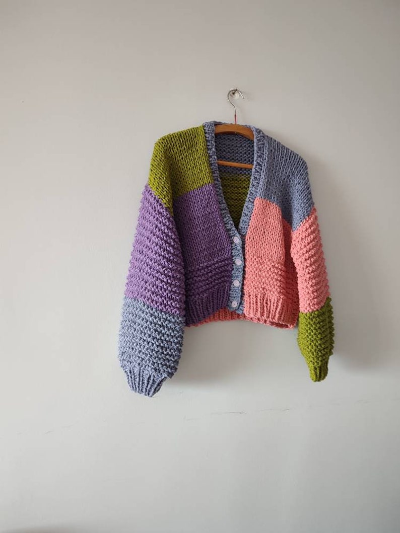 Op de afbeelding: Een chunky gebreid vest in een patchwork-ontwerp, met panelen in lavendel, lichtblauw, roze, groen en olijfgroen. De cropped trui heeft een V-hals, knoopsluiting en lange mouwen. Het hangt aan een houten hanger tegen een witte muur.