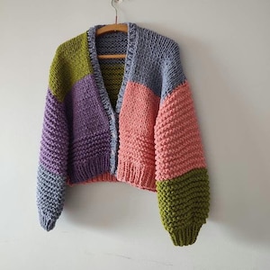 Op de afbeelding: Een grofgebreid vest in een patchwork-ontwerp, met panelen in paars, olijfgroen, roze en blauw. De open trui hangt aan een houten hanger tegen een witte muur. De mouwen zijn lang en de pasvorm is relaxed.
