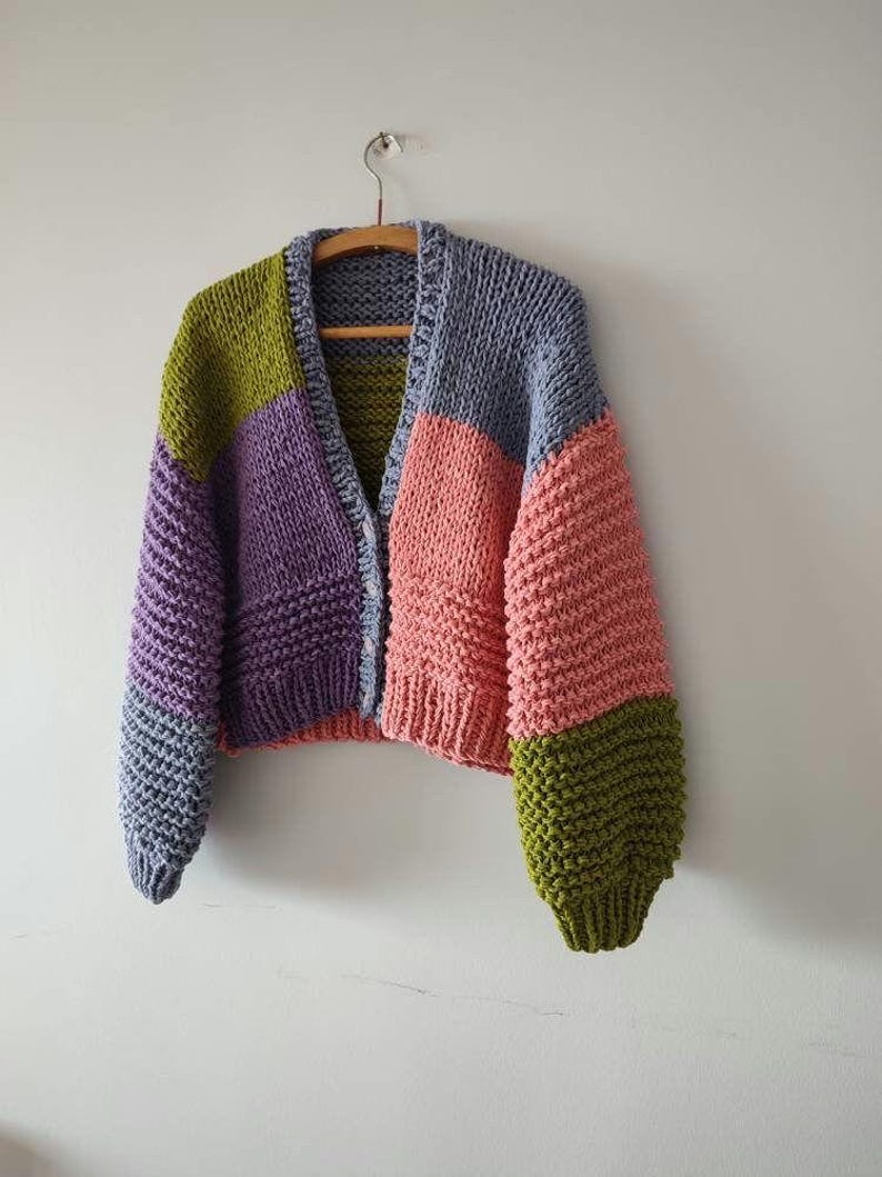 Op de afbeelding: Een grofgebreid vest in een patchwork-ontwerp, met panelen in paars, olijfgroen, roze en blauw. De open trui hangt aan een houten hanger tegen een witte muur. De mouwen zijn lang en de pasvorm is relaxed.