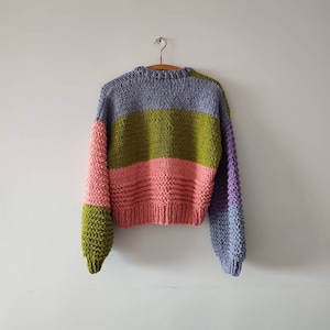 Op de afbeelding: Een handgebreide trui met kleurvlakken in lavendel, olijfgroen, roze en paars. De trui hangt aan een houten kleerhanger tegen een witte muur. De mouwen hebben een andere kleur dan de body.