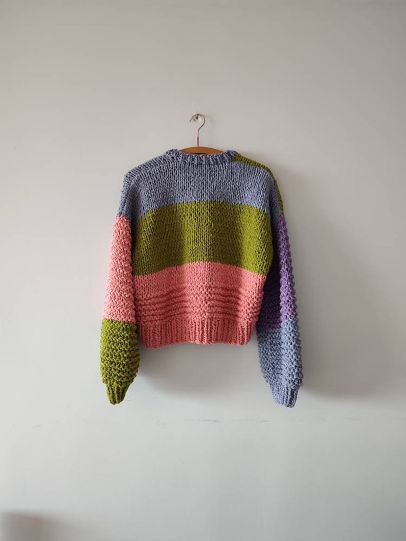 Op de afbeelding: Een handgebreide trui met kleurvlakken in lavendel, olijfgroen, roze en paars. De trui hangt aan een houten kleerhanger tegen een witte muur. De mouwen hebben een andere kleur dan de body.