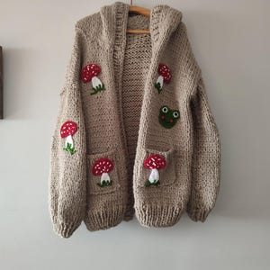 May include: A beige knitted cardigan with a hood, featuring embroidered mushrooms and a frog. The cardigan has two pockets with additional mushroom embroidery.