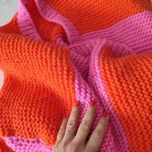 May include: A hand-knitted tote bag featuring a vibrant color-blocked pattern of bright orange and hot pink yarn. The bag is constructed with a textured garter stitch and includes two integrated shoulder straps in a matching pink hue.