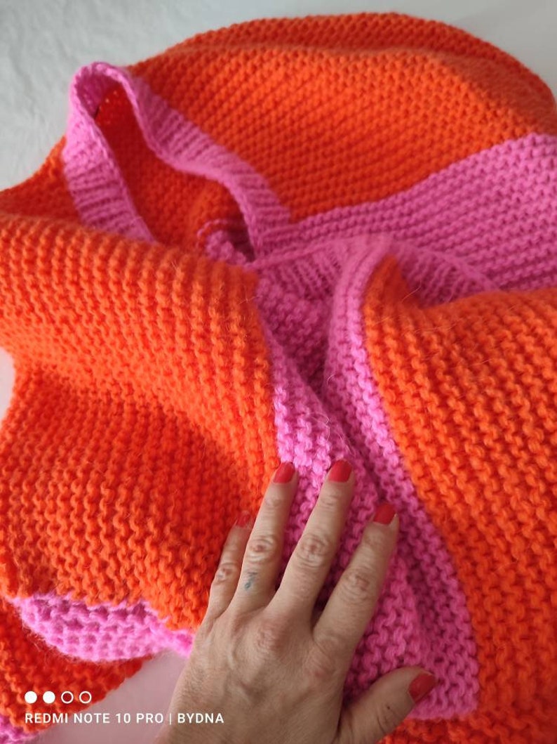 May include: A hand-knitted tote bag featuring a vibrant color-blocked pattern of bright orange and hot pink yarn. The bag is constructed with a textured garter stitch and includes two integrated shoulder straps in a matching pink hue.