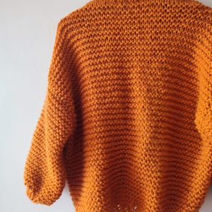 Puede incluir: Un su&eacute;ter de punto grueso en un vibrante color naranja quemado. La prenda presenta una textura de punto abierto y suelto con mangas largas y un cuello acanalado, colgado de una percha sobre un fondo liso de color claro.