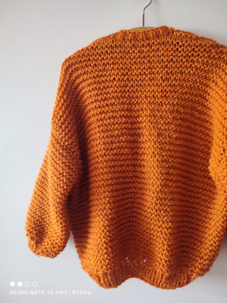Puede incluir: Un su&eacute;ter de punto grueso en un vibrante color naranja quemado. La prenda presenta una textura de punto abierto y suelto con mangas largas y un cuello acanalado, colgado de una percha sobre un fondo liso de color claro.