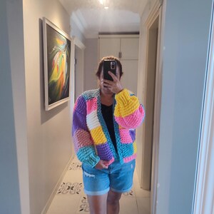 May include: A colorful, chunky-knit cardigan in a patchwork design. The sweater features blocks of purple, yellow, pink, blue, and white. The sleeves are puffy, and the cardigan is open in the front. The person is wearing denim shorts.