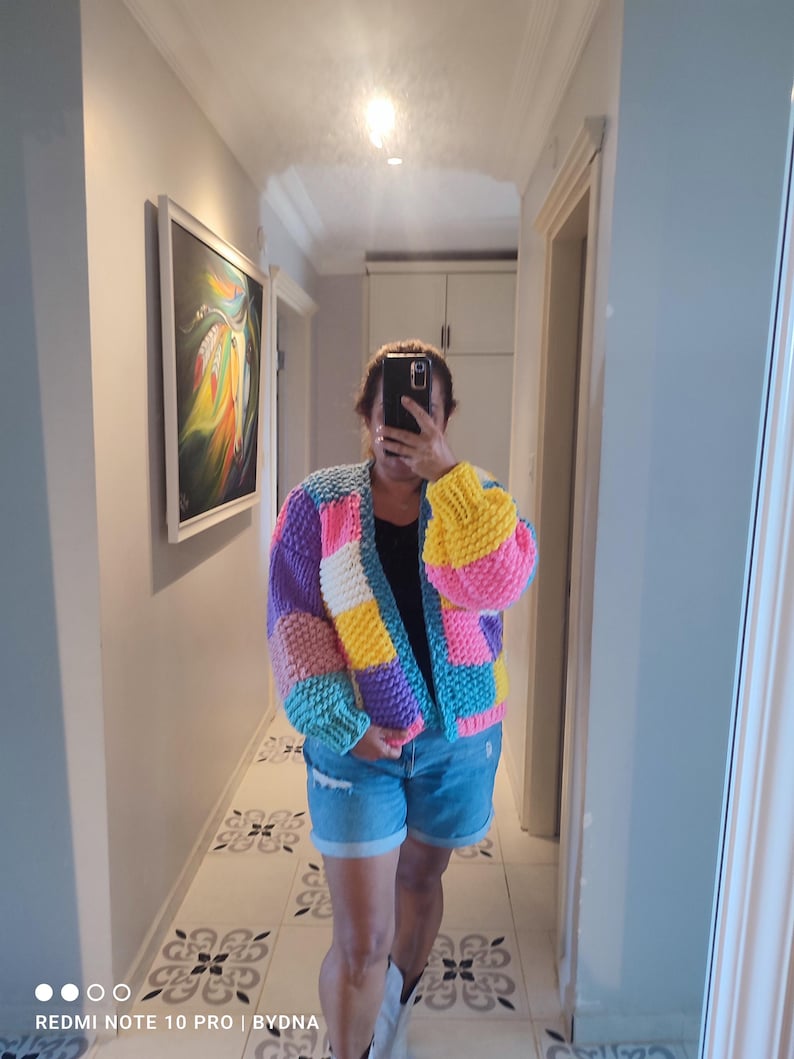 May include: A colorful, chunky-knit cardigan in a patchwork design. The sweater features blocks of purple, yellow, pink, blue, and white. The sleeves are puffy, and the cardigan is open in the front. The person is wearing denim shorts.