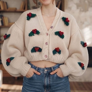 May include: A cream-colored knit cardigan featuring a V-neckline, long balloon sleeves, and a button-down front. The garment is decorated with multiple small, three-dimensional crocheted holly motifs, each consisting of dark green leaves and red berries.