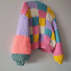 May include: A colorful, patchwork-style knitted sweater. The sweater features a variety of pastel colors, including pink, yellow, blue, and lavender. The sleeves are a contrasting coral color with a teal cuff. The sweater is hanging on a hook.