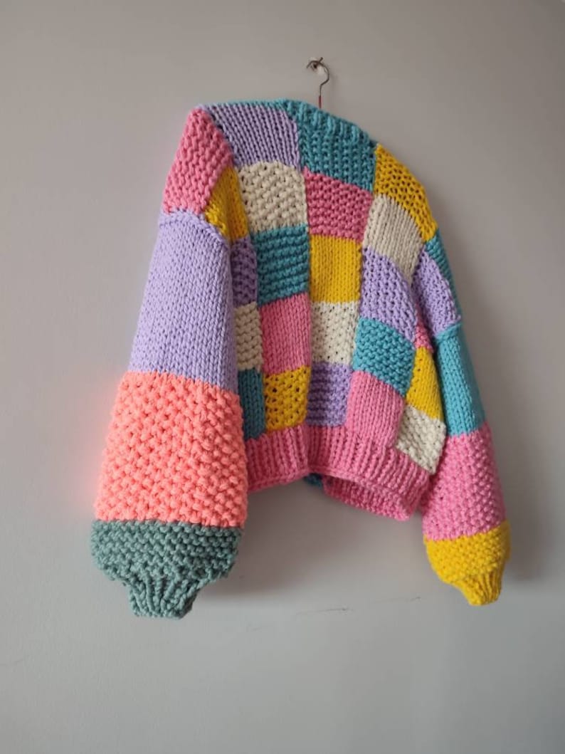 May include: A colorful, patchwork-style knitted sweater. The sweater features a variety of pastel colors, including pink, yellow, blue, and lavender. The sleeves are a contrasting coral color with a teal cuff. The sweater is hanging on a hook.