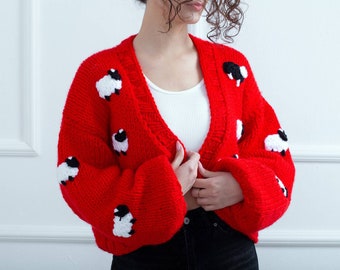 Könnte beinhalten: Ein leuchtend roter, grob gestrickter Cardigan mit Schafmuster. Die Schafe sind weiß mit schwarzen Gesichtern und gleichmäßig über dem Pullover verteilt. Der Cardigan hat einen V-Ausschnitt und lange Ärmel.