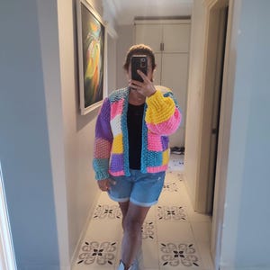 May include: A colorful, patchwork knit cardigan in shades of blue, pink, yellow, purple, and white. The open-front sweater has a relaxed fit and long sleeves. The person in the image is wearing denim shorts and white boots.