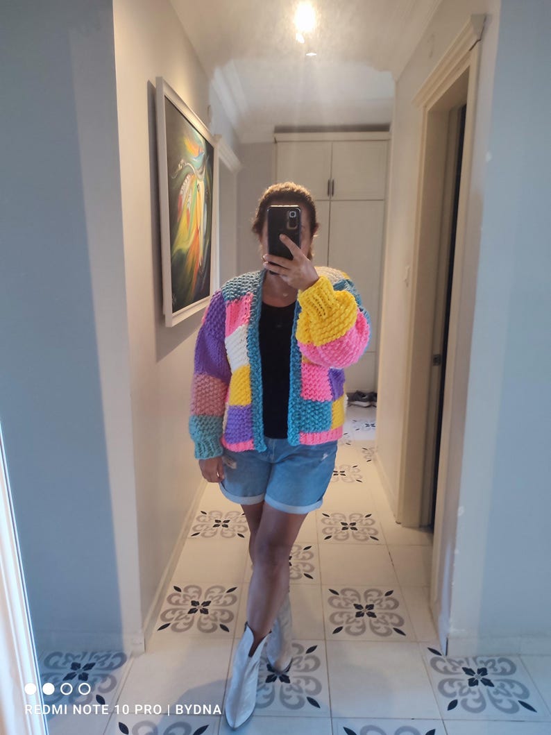 May include: A colorful, patchwork knit cardigan in shades of blue, pink, yellow, purple, and white. The open-front sweater has a relaxed fit and long sleeves. The person in the image is wearing denim shorts and white boots.