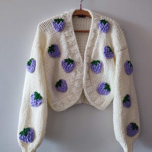 May include: A cream-coloured, open-front cardigan with purple and green berry appliqués. The sweater has long sleeves and a V-neck design, hanging on a wooden hanger against a plain white wall.