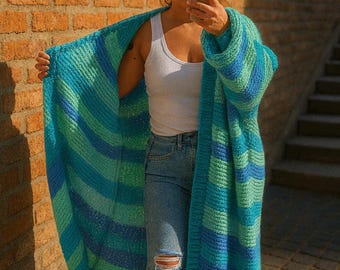 May include: A long, oversized cardigan in shades of blue and green stripes. The open-front sweater is made of a chunky knit material. The person is wearing a white tank top and blue jeans. The background is a brick wall.