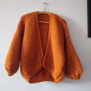 Puede incluir: Un c&aacute;rdigan de punto grueso en un vibrante color naranja quemado. Esta prenda abierta en la parte delantera presenta mangas largas y voluminosas con pu&ntilde;os acanalados y un patr&oacute;n de punto bobo texturizado en todo el cuerpo, colgado en una percha de madera.