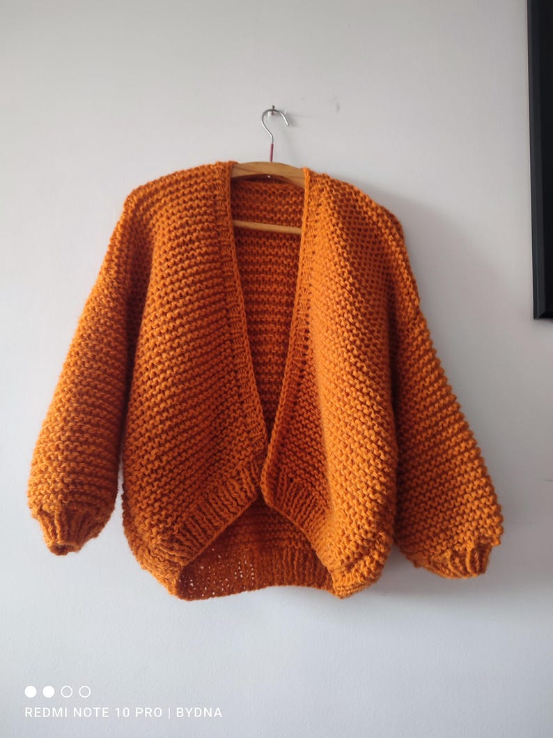 Puede incluir: Un c&aacute;rdigan de punto grueso en un vibrante color naranja quemado. Esta prenda abierta en la parte delantera presenta mangas largas y voluminosas con pu&ntilde;os acanalados y un patr&oacute;n de punto bobo texturizado en todo el cuerpo, colgado en una percha de madera.