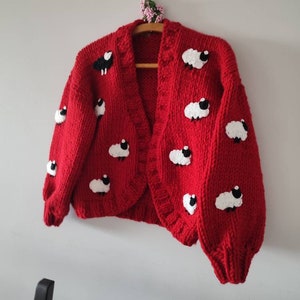 K&ouml;nnte beinhalten: Ein leuchtend roter Cardigan, verziert mit wei&szlig;en und schwarzen Schafmotiven. Der offene Pullover wird auf einem Holzb&uuml;gel pr&auml;sentiert. Der grob gestrickte Pullover hat einen runden Ausschnitt und lange &Auml;rmel.