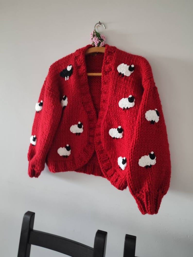 K&ouml;nnte beinhalten: Ein leuchtend roter Cardigan, verziert mit wei&szlig;en und schwarzen Schafmotiven. Der offene Pullover wird auf einem Holzb&uuml;gel pr&auml;sentiert. Der grob gestrickte Pullover hat einen runden Ausschnitt und lange &Auml;rmel.