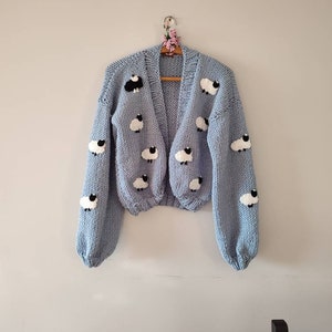 K&ouml;nnte beinhalten: Hellblauer Cardigan mit V-Ausschnitt und langen &Auml;rmeln, verziert mit wei&szlig;en Schafen mit schwarzen Gesichtern. Der Strickpullover h&auml;ngt an einem Holzkleiderb&uuml;gel. Das Kleidungsst&uuml;ck wird vor einer schlichten Wand pr&auml;sentiert.