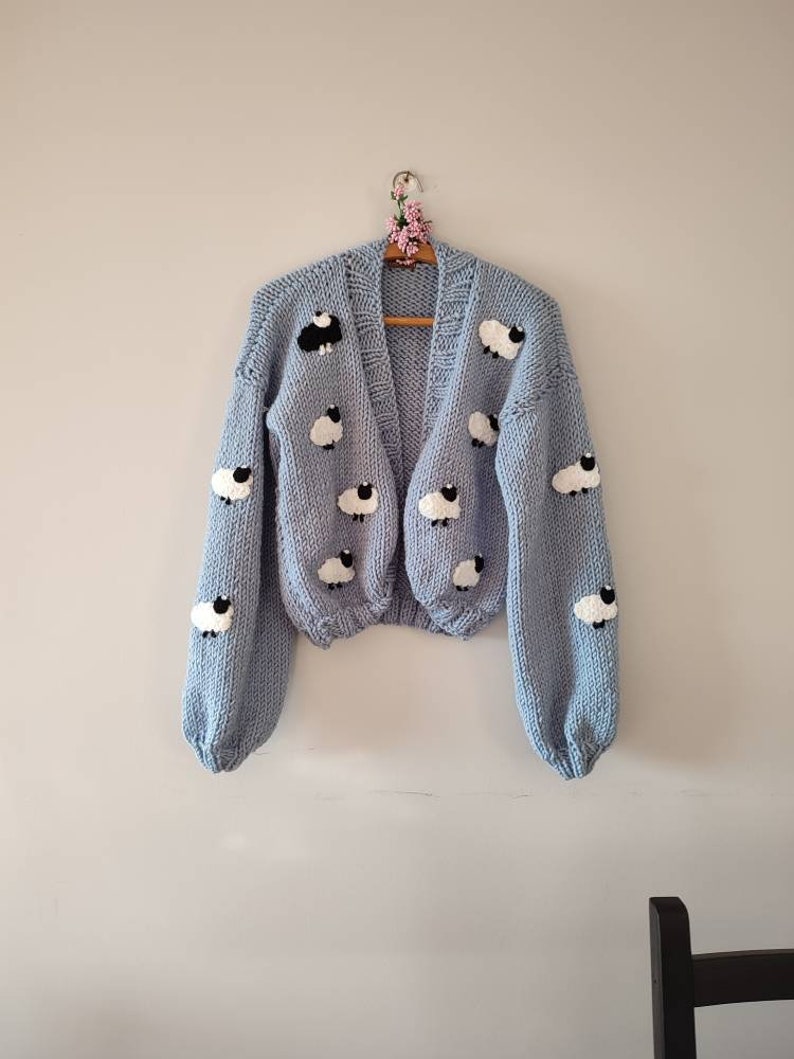 K&ouml;nnte beinhalten: Hellblauer Cardigan mit V-Ausschnitt und langen &Auml;rmeln, verziert mit wei&szlig;en Schafen mit schwarzen Gesichtern. Der Strickpullover h&auml;ngt an einem Holzkleiderb&uuml;gel. Das Kleidungsst&uuml;ck wird vor einer schlichten Wand pr&auml;sentiert.