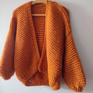 Puede incluir: Un c&aacute;rdigan de punto grueso en un vibrante color naranja quemado. Esta prenda abierta por delante presenta mangas largas y voluminosas con pu&ntilde;os acanalados y una textura de hilo grueso. El c&aacute;rdigan se muestra colgado en una percha de madera.