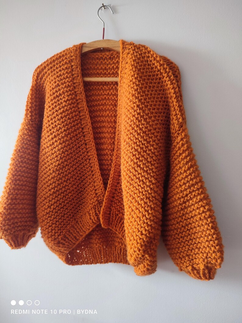 Puede incluir: Un c&aacute;rdigan de punto grueso en un vibrante color naranja quemado. Esta prenda abierta por delante presenta mangas largas y voluminosas con pu&ntilde;os acanalados y una textura de hilo grueso. El c&aacute;rdigan se muestra colgado en una percha de madera.