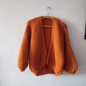 Puede incluir: Un c&aacute;rdigan de punto grueso en un vibrante color naranja quemado. Esta prenda abierta en la parte delantera presenta mangas largas con pu&ntilde;os acanalados y un patr&oacute;n de punto texturizado en toda la pieza. El c&aacute;rdigan cuelga de una percha de madera.