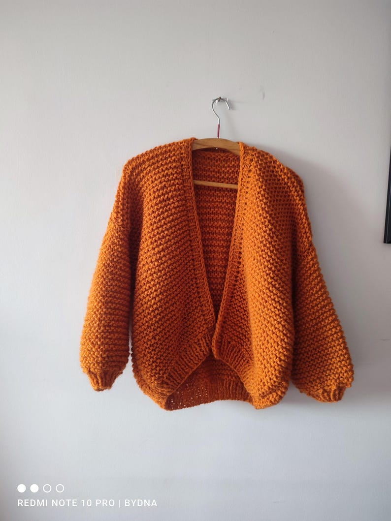 Puede incluir: Un c&aacute;rdigan de punto grueso en un vibrante color naranja quemado. Esta prenda abierta en la parte delantera presenta mangas largas con pu&ntilde;os acanalados y un patr&oacute;n de punto texturizado en toda la pieza. El c&aacute;rdigan cuelga de una percha de madera.