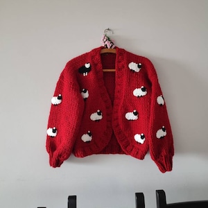 K&ouml;nnte beinhalten: Ein roter Cardigan mit wei&szlig;en und schwarzen Schafmotiven h&auml;ngt an einem Holzb&uuml;gel. Der offene Pullover hat ein grobes Strickdesign. Das Kleidungsst&uuml;ck wird vor einer schlichten wei&szlig;en Wand pr&auml;sentiert.