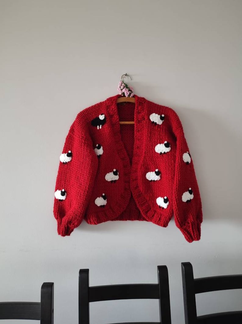 K&ouml;nnte beinhalten: Ein roter Cardigan mit wei&szlig;en und schwarzen Schafmotiven h&auml;ngt an einem Holzb&uuml;gel. Der offene Pullover hat ein grobes Strickdesign. Das Kleidungsst&uuml;ck wird vor einer schlichten wei&szlig;en Wand pr&auml;sentiert.