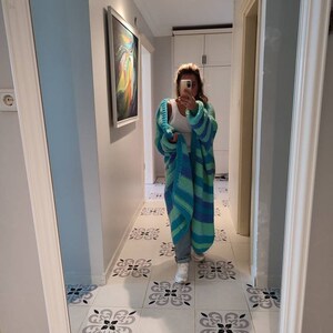 May include: A long, open-front cardigan in shades of blue and green. The chunky knitwear features wide stripes and a relaxed fit. The cardigan is worn over a white top and jeans. The image is taken in a hallway with patterned tile flooring.
