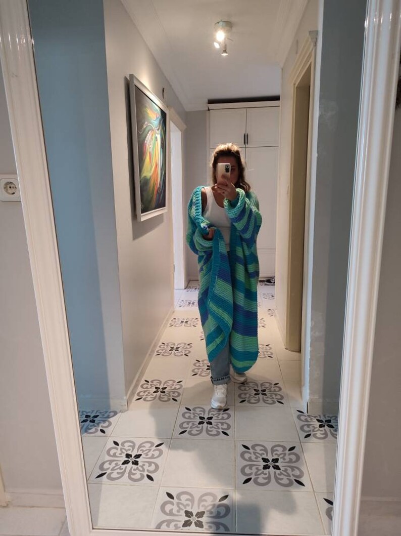 May include: A long, open-front cardigan in shades of blue and green. The chunky knitwear features wide stripes and a relaxed fit. The cardigan is worn over a white top and jeans. The image is taken in a hallway with patterned tile flooring.