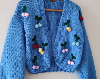 May include: A light blue, open-front cardigan sweater with a chunky knit texture. The sweater is adorned with colorful, three-dimensional cherry appliqués in red, pink, yellow, and blue, each with green leaves. The sweater is hanging on a wooden hanger.