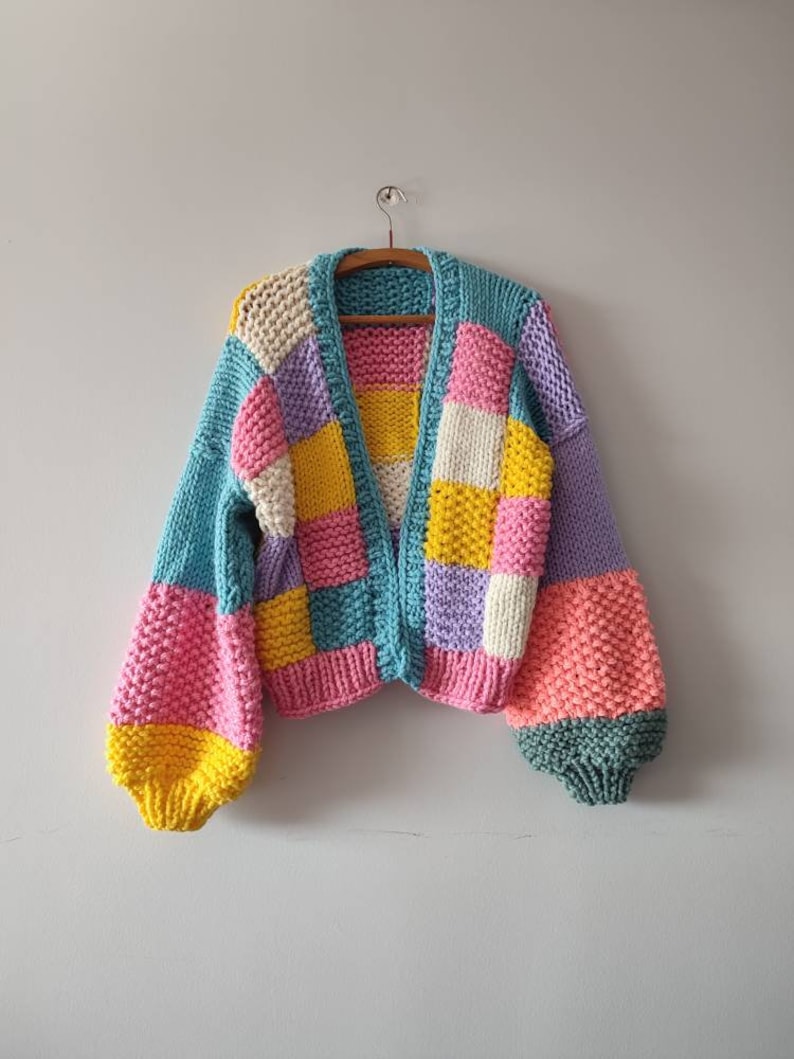 May include: A colorful, patchwork-style cardigan in shades of pink, yellow, blue, white, and purple. The knitted sweater features a V-neck and long, puffy sleeves. The garment is hanging on a wooden hanger against a plain white wall.