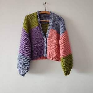Op de afbeelding: Een chunky gebreid vest in een colorblock-ontwerp. De trui heeft een V-hals en een knoopsluiting. De kleurblokken omvatten tinten paars, roze, groen en blauw. Het vest hangt aan een houten hanger tegen een witte muur.