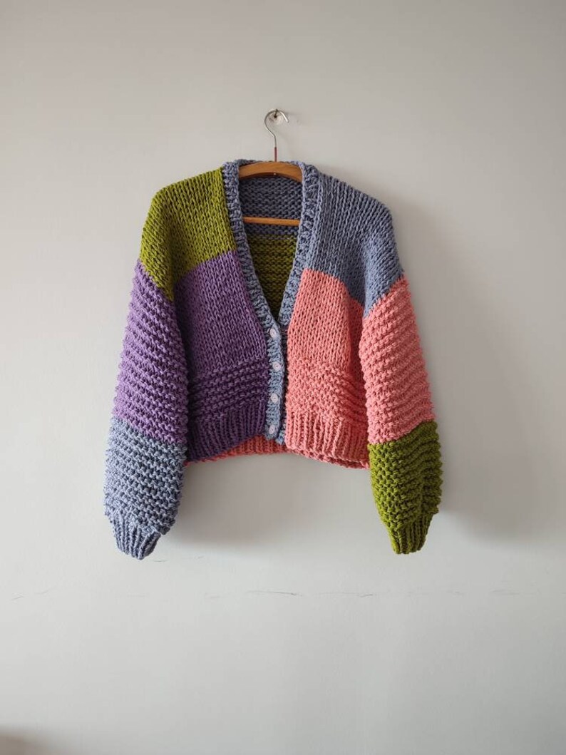 Op de afbeelding: Een chunky gebreid vest in een colorblock-ontwerp. De trui heeft een V-hals en een knoopsluiting. De kleurblokken omvatten tinten paars, roze, groen en blauw. Het vest hangt aan een houten hanger tegen een witte muur.