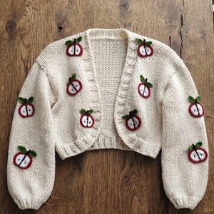 Puede incluir: Un cárdigan de punto color crema con mangas largas y corte corto. La prenda está decorada con múltiples motivos de manzanas de ganchillo en rojo, blanco y verde, distribuidos por los paneles delanteros y las mangas.