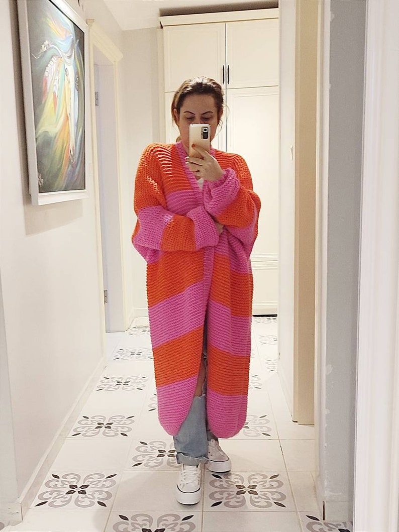 May include: A long, chunky knit cardigan featuring horizontal stripes in vibrant orange and bright pink. The garment has an open front, long sleeves, and a thick, textured stitch pattern. It is paired with light blue denim jeans and white sneakers.