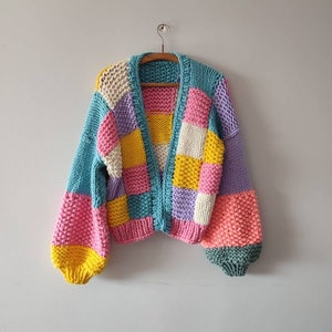 May include: A colorful, patchwork-style cardigan in shades of blue, pink, yellow, and purple. The open-front sweater features a chunky knit design with long, puffy sleeves. The garment is displayed on a wooden hanger against a plain wall.