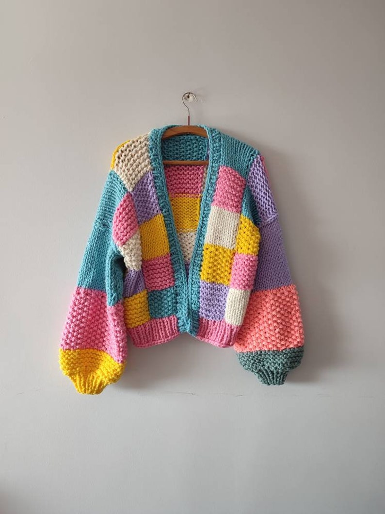 May include: A colorful, patchwork-style cardigan in shades of blue, pink, yellow, and purple. The open-front sweater features a chunky knit design with long, puffy sleeves. The garment is displayed on a wooden hanger against a plain wall.