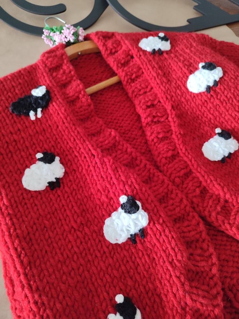 K&ouml;nnte beinhalten: Ein leuchtend roter Strickcardigan, verziert mit wei&szlig;en und schwarzen Schafapplikationen. Der Pullover hat ein strukturiertes Strickmuster und wird auf einem Holzkleiderb&uuml;gel pr&auml;sentiert. Die Schafe sind gleichm&auml;&szlig;ig &uuml;ber die Vorderseite des Cardigans verteilt.
