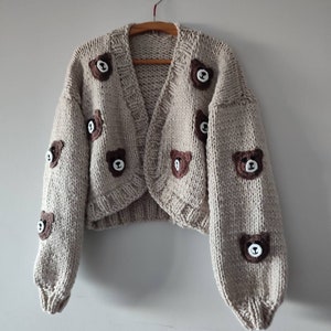 May include: A beige chunky knit cardigan featuring long sleeves and a V-neckline. The garment is decorated with multiple small, dark brown crocheted bear face appliques scattered across the front and sleeves, each with white stitched details.