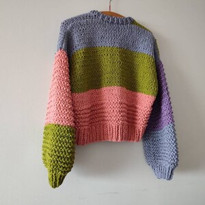 Op de afbeelding: Een chunky gebreide trui in een colorblock-ontwerp. De trui heeft een combinatie van kleuren, waaronder lavendel, olijfgroen, roze en paars. De mouwen hebben een andere kleur dan de body. De trui hangt aan een houten hanger.