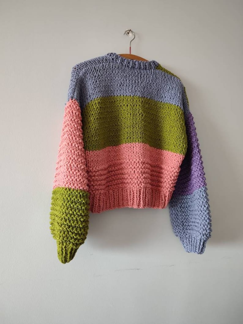 Op de afbeelding: Een chunky gebreide trui in een colorblock-ontwerp. De trui heeft een combinatie van kleuren, waaronder lavendel, olijfgroen, roze en paars. De mouwen hebben een andere kleur dan de body. De trui hangt aan een houten hanger.