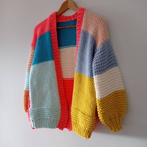 Puede incluir: Un cárdigan de punto grueso en un mosaico de colores, incluyendo coral, turquesa, azul claro, blanco, amarillo y melocotón. El suéter abierto cuelga de una percha de madera. Las mangas son abullonadas.