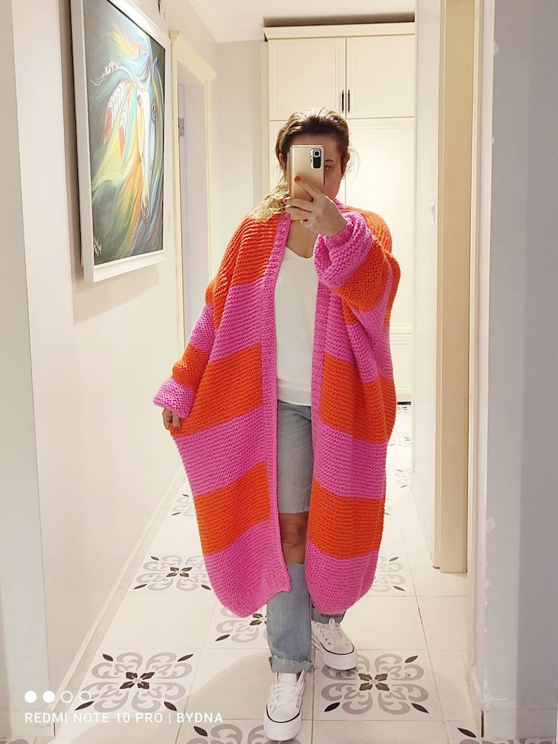 May include: A long, chunky knit cardigan featuring wide horizontal stripes in vibrant orange and bright pink. The garment has an open front, long sleeves, and a thick, textured stitch pattern that extends to the mid-calf length of the piece.