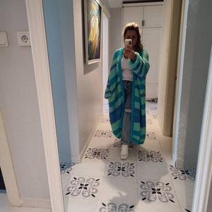 May include: A long, open-front cardigan in shades of blue and green, with horizontal stripes. The cardigan is being worn in a hallway with a decorative tile floor and a large mirror.